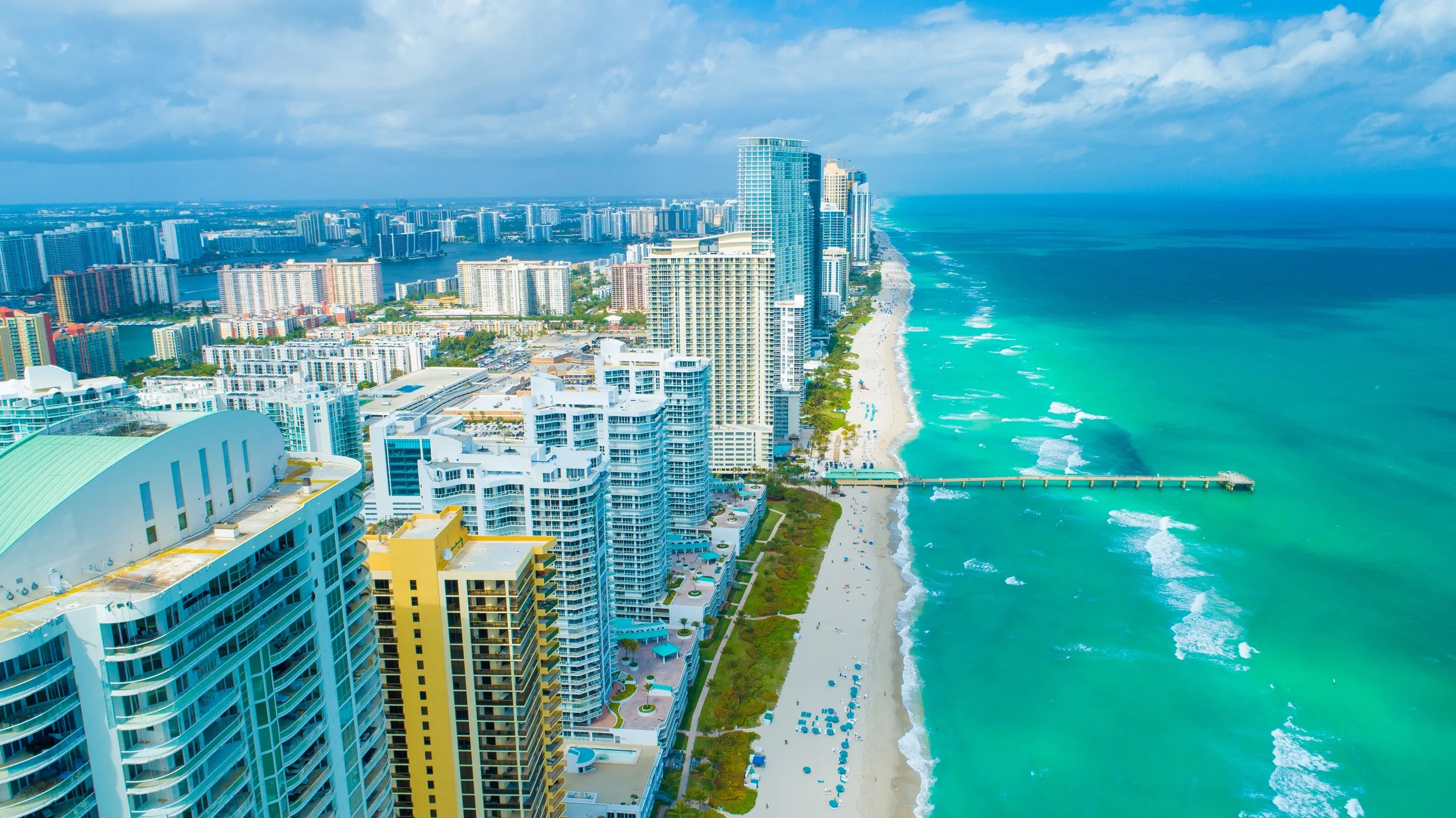 Aerial view along the Sunny Isles Beach oceanfront, towers lining the sand with the fishing pier beyond
